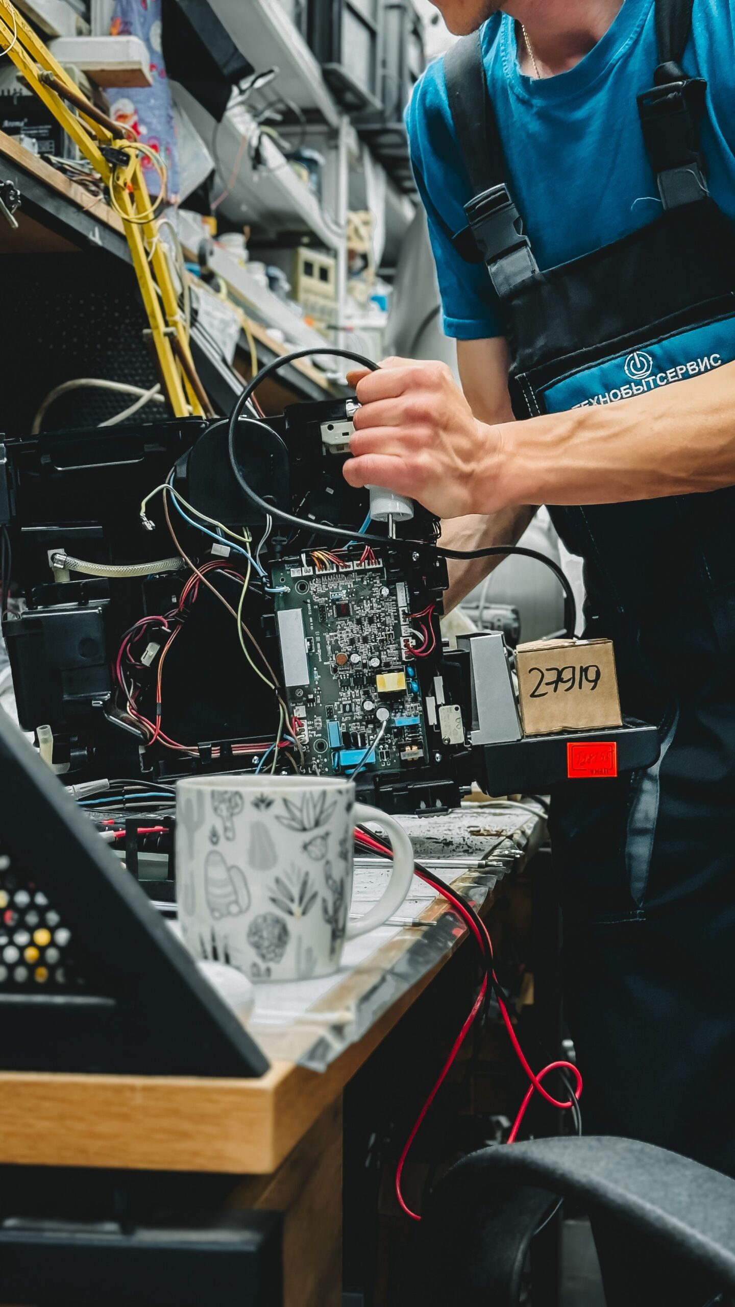 Technician working on printer repair with tools in a cluttered workshop environment.